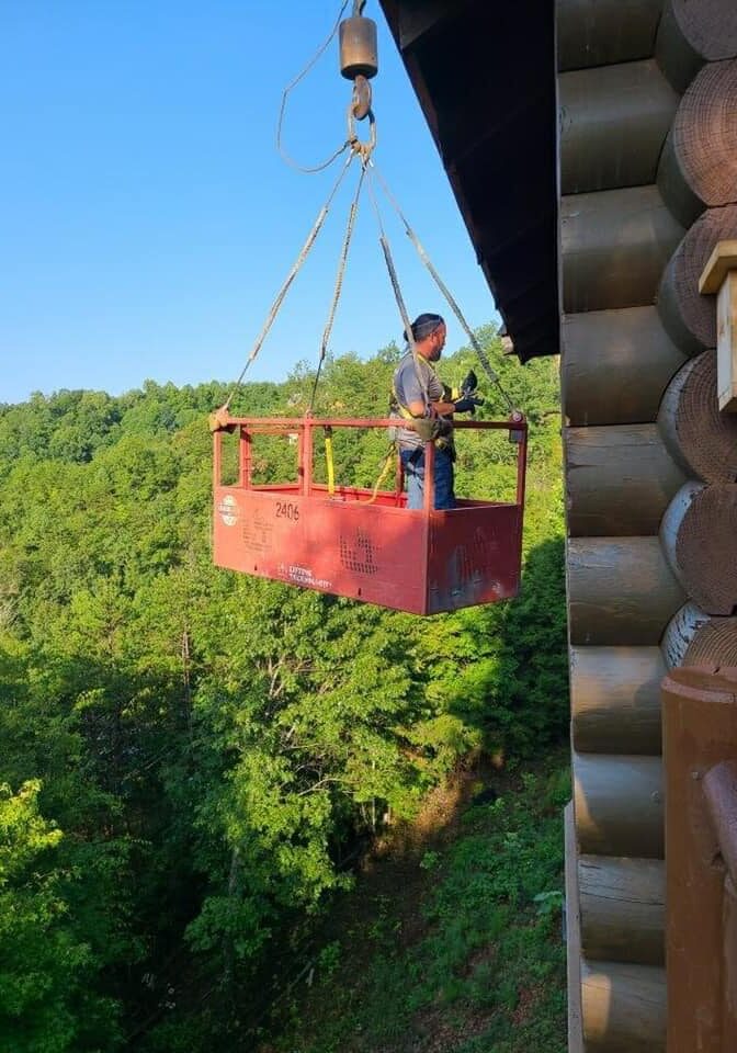 A person in a red basket lifted by a crane above green trees.